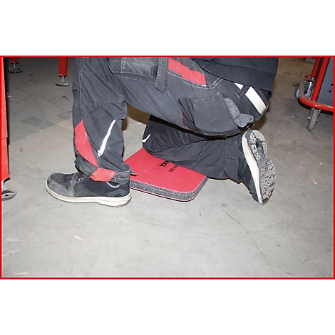 Arbeiter kniet auf einer roten Knieschutzmatte, schwarze Hose mit roten Akzenten, schwarze Schuhe.